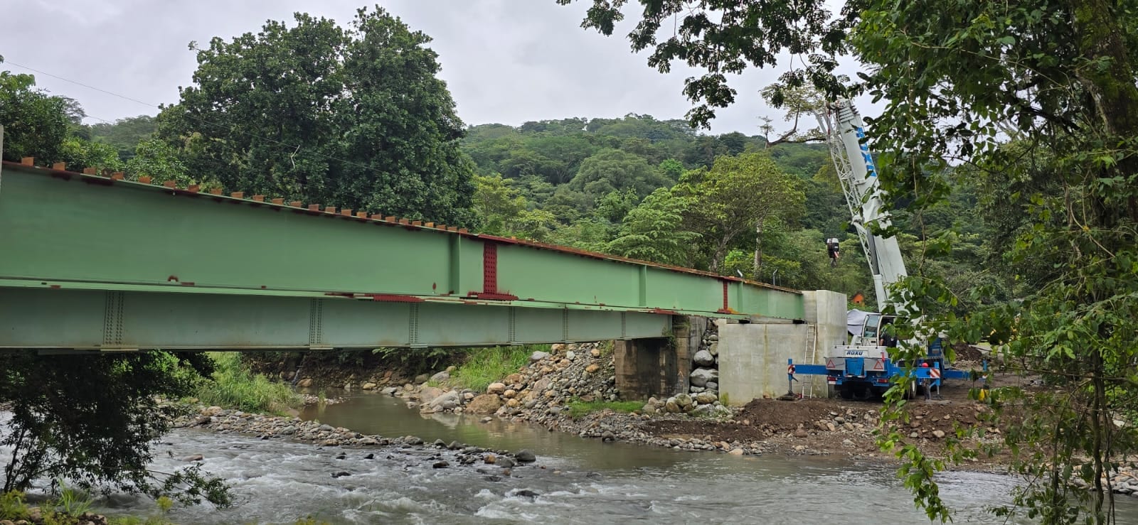 Puente de vigas metálicas verdes sobre un río, con una grúa al fondo.